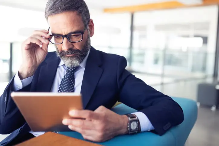 A man with glasses and a gray beard sits in an office environment, holding a tablet and adjusting his glasses with his other hand. He's wearing a suit and appears focused.