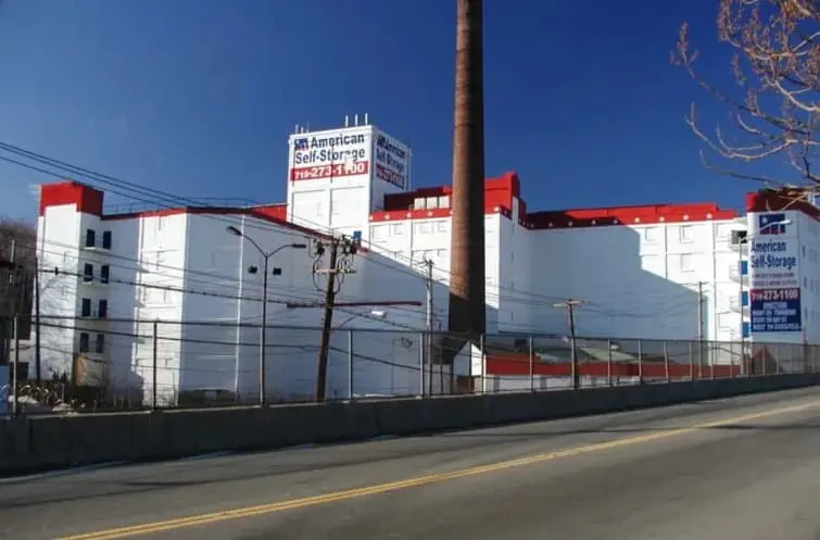 Large white storage facility with red accents, labeled "American Self Storage," adjacent to a tall chimney. A road and chain-link fence are in the foreground under a clear blue sky.