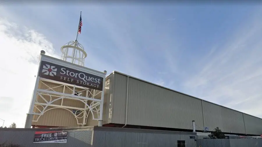 A large StorQuest Self Storage facility with an American flag on top, featuring a high structure at the corner. The exterior is beige with a prominent sign and a banner offering free truck rental.