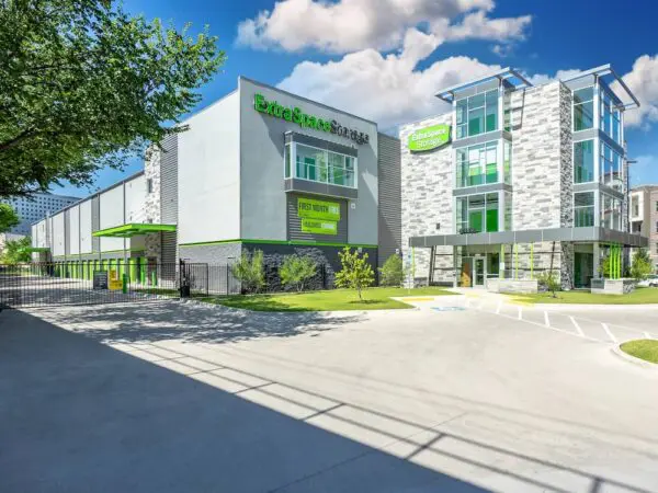 Modern storage facility with green accents, featuring clear signage and multiple stories. The building is surrounded by trees and a paved parking area under a partly cloudy sky.
