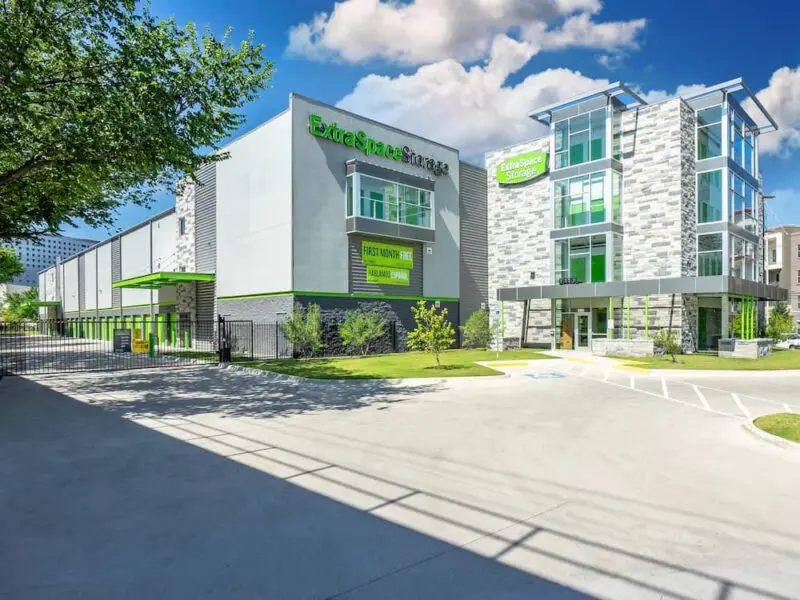 Modern storage facility with green accents, featuring clear signage and multiple stories. The building is surrounded by trees and a paved parking area under a partly cloudy sky.