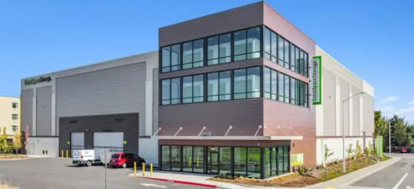 A modern three-story building with large windows, labeled "Life Storage," stands next to a large warehouse. Several small vehicles are parked outside under a clear blue sky.