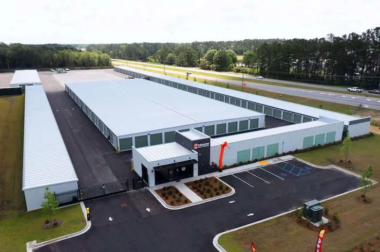 Aerial view of a large storage facility with numerous units and a parking area, surrounded by trees and a road.