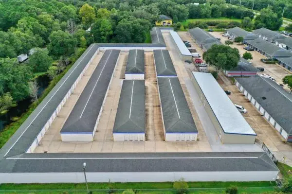 Aerial view of a large self-storage facility featuring multiple long, rectangular storage units surrounded by trees and neighboring buildings.