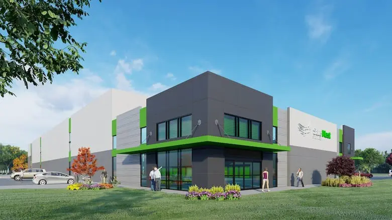Modern industrial building with a green and gray exterior, featuring large windows. People are walking nearby, and a few cars are parked in the lot. Clear blue sky in the background.