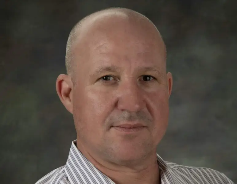 A bald man wearing a striped shirt poses against a dark, textured background.