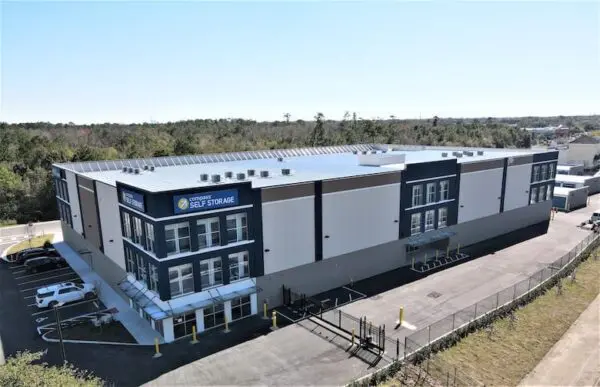Aerial view of a large, rectangular self-storage facility with a parking lot, surrounded by trees and fencing. The building is modern with white and dark blue accents.