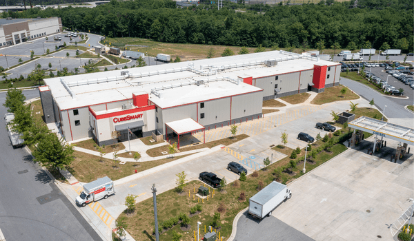 Aerial view of a storage facility with white and red buildings, parking lot, and surrounding greenery on a clear day.