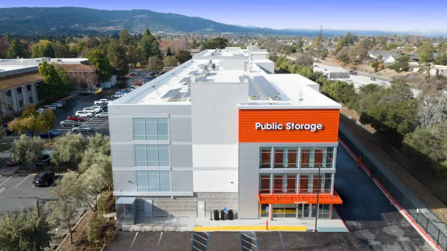 Aerial view of a multi-story Public Storage building with solar panels on the roof, surrounded by a parking lot and green landscape.