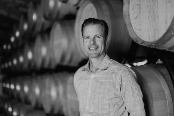 A person in a plaid shirt stands smiling in front of stacked wooden barrels in a dimly lit room.