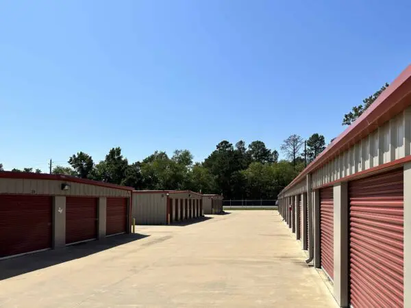 A row of storage units with red rolling doors on a sunny day, surrounded by trees and a clear blue sky.