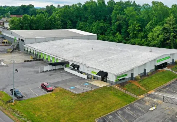 Large white building with flat roofs surrounded by trees, featuring parking spaces and a single red car. A green sign reads "Store.