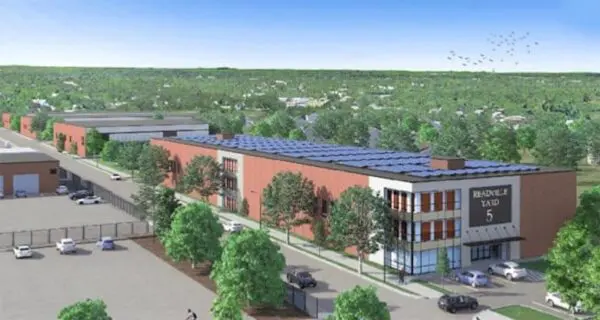 Aerial view of an industrial complex with solar panels on the roof, surrounded by trees, parking lot, and distant residential homes under a clear blue sky.