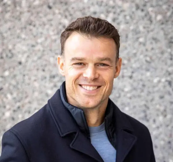 Man in a black coat smiling in front of a speckled gray wall.