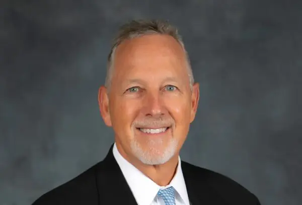 Man in a suit with a light blue tie, smiling against a dark gray backdrop.