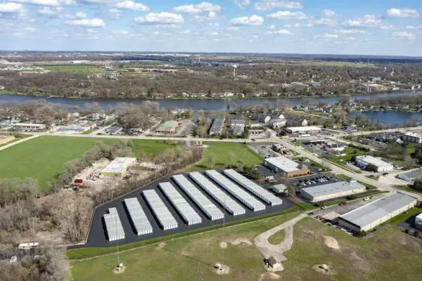 Aerial view of a storage facility with multiple rows of units, surrounded by green fields and near a river, with scattered residential and industrial buildings in the background under a blue sky.