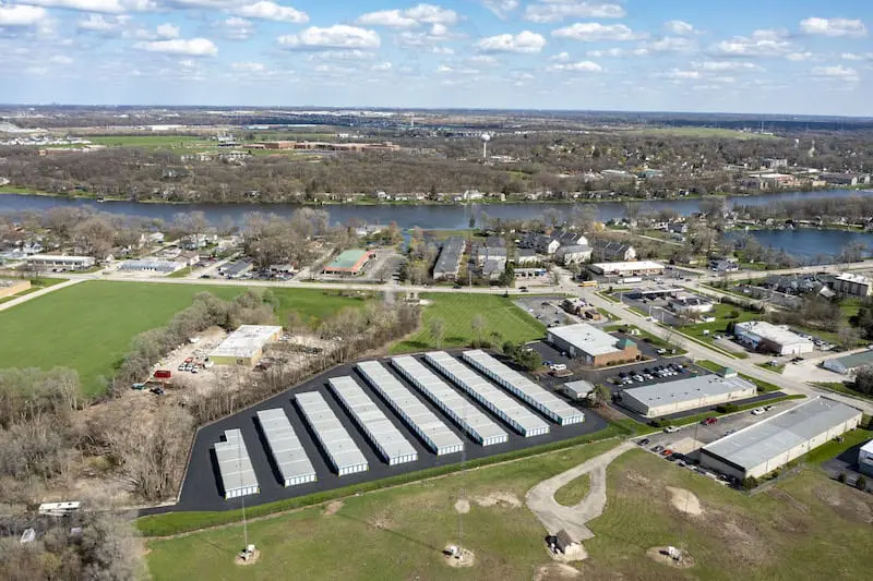 Aerial view of a storage facility with multiple rows of units, surrounded by green fields and near a river, with scattered residential and industrial buildings in the background under a blue sky.