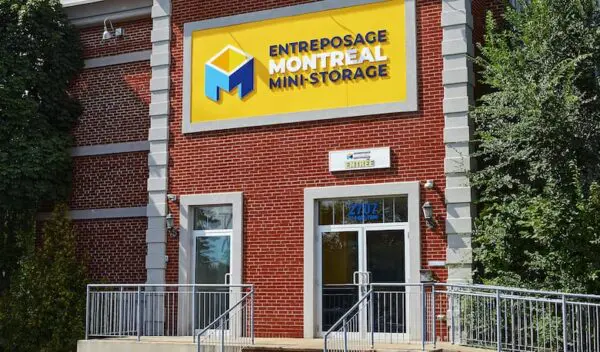 Entrance to Entrepôt Montréal Mini-Storage with a yellow sign and glass doors, surrounded by brick walls and greenery.