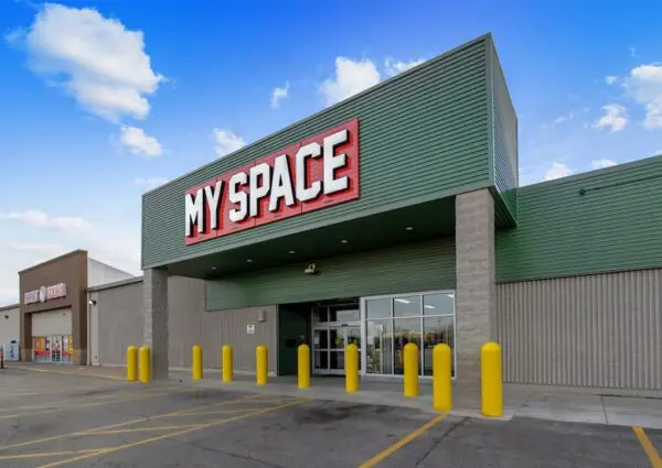 A retail store entrance with a green and gray facade features large red and white "MY SPACE" signage. Bright yellow bollards line the entrance area.