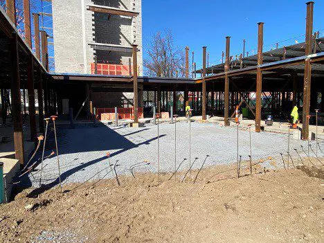 Construction site with metal structures and a gravel-covered ground. Workers in safety gear are visible. A building and a clear blue sky are in the background.