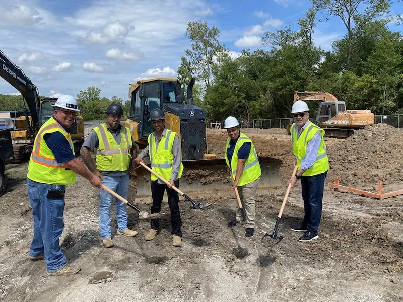 Five people in safety vests and helmets stand holding shovels at a construction site with excavators and a bulldozer in the background.