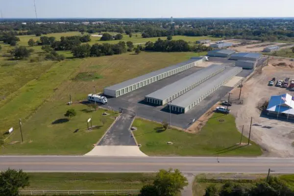 Aerial view of a storage facility with multiple long, white-roofed buildings in a rural area, surrounded by fields and a few trees. A road runs in front of the facility.