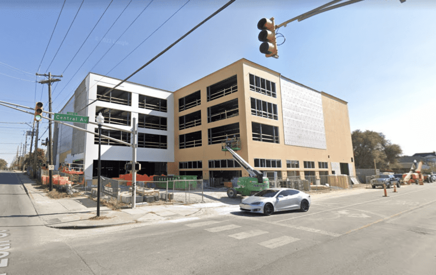 A white four-story building under construction at a street intersection with a traffic light and a parked white car in the foreground.