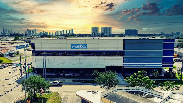 Large, modern building with blue accents and "snapbox" signage, situated on a street corner, with a city skyline visible in the background under a partly cloudy sky.