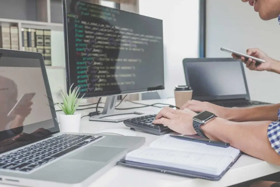 A person is typing on their computer, which has two screens and codes on it.