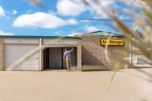 A person stands in front of a row of self-storage units, opening the door to unit 229. Blue sky and grass in the foreground.