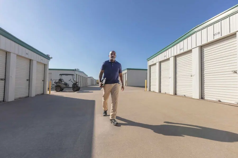 A man walks on a concrete path between storage units under a clear blue sky, carrying a clipboard. A golf cart is parked on the left.