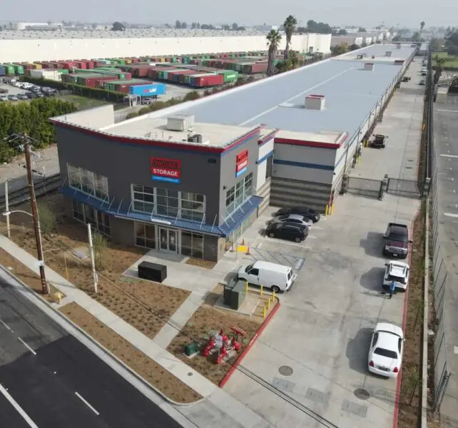 Aerial view of a self-storage facility with a large warehouse, a parking area, and several parked vehicles on a clear day. Rows of shipping containers are visible in the background.