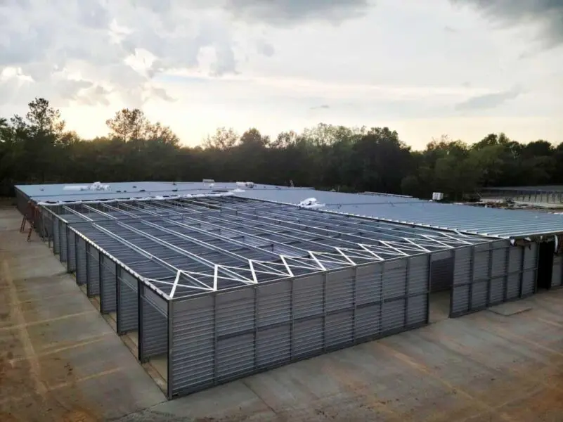 Large unfinished steel structure with open roof beams and walls, surrounded by trees under a cloudy sky.