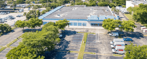 Aerial view of a large commercial building surrounded by a mostly empty parking lot with several trees. A few vehicles are parked on one side.