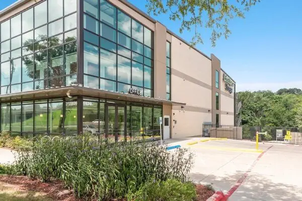 Modern two-story office building with large windows, surrounded by plants and a clear parking area. The sky is clear and blue.