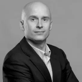 A bald man in a suit and checkered shirt poses against a gray background, looking directly at the camera with a neutral expression.