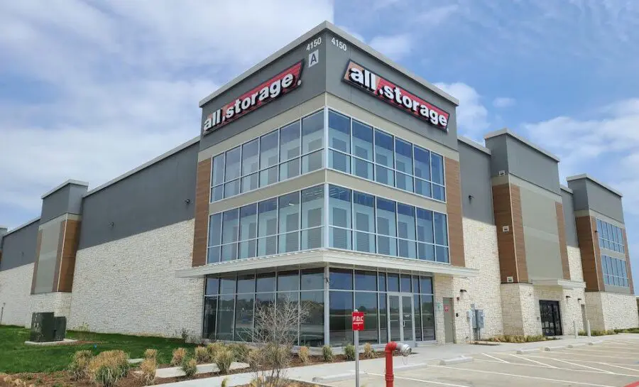Modern self-storage facility with multiple floors, large windows, and a "storage" sign. The building is surrounded by a parking area and landscaped grounds.