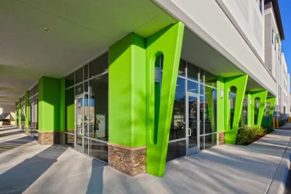 Modern building exterior with bright green walls, large glass windows, and stone accents. The sidewalk is visible in the foreground.