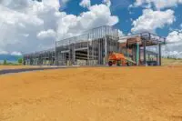 A metal-framed building under construction stands on a dirt lot, with an orange lift parked nearby and clouds in the sky above.