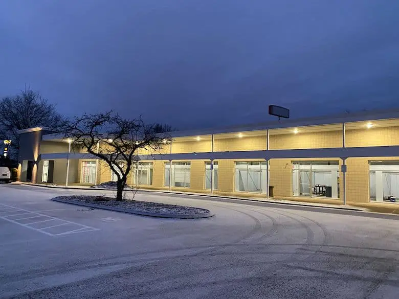 Two-story building with illuminated windows at dusk. Empty parking lot with a tree and subtle snowfall on the ground.