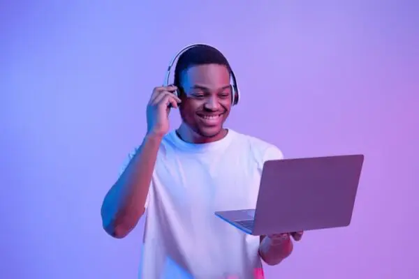 Man with headphones smiling at a laptop in left hand, holding a phone to his ear with right hand, against a blue and purple gradient background.