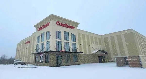 Large beige building with "CubeSmart" signage, surrounded by snow. A single car is parked to the side. Overcast sky with falling snowflakes.