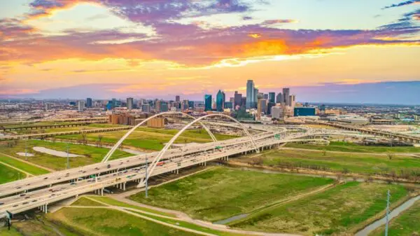 Aerial view of a city skyline during sunset, featuring a prominent bridge with multiple arches and surrounding highways set against a colorful sky.
