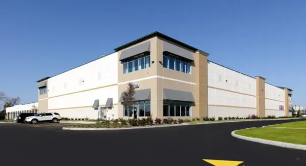 Large warehouse building with beige and white exterior, parking area with cars, and landscaped greenery under a clear blue sky.