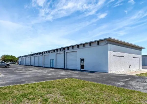 Long white warehouse building with multiple garage doors, situated on a paved lot with a grassy area in the foreground. A car is parked to the left. Blue sky with light clouds above.