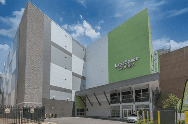 Exterior of an Extra Space Storage facility with a green and gray facade, a fenced entrance, and a clear blue sky in the background.
