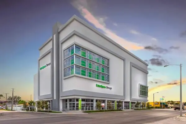Modern storage facility building with green and white accents, large windows, and a flat roof, located at a street corner under a blue sky with clouds.