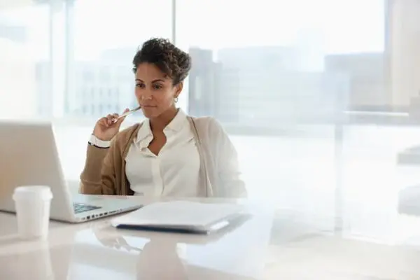 A person sits at a desk, looking at a laptop, holding a pen. Papers and a takeaway coffee cup are on the desk. Bright, modern office setting with large windows in the background.
