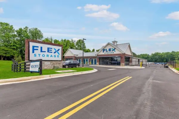 Flex Storage facility entrance with signage and paved road. A single-story building with a metal roof, surrounded by trees under a blue sky. A car is parked near the front.