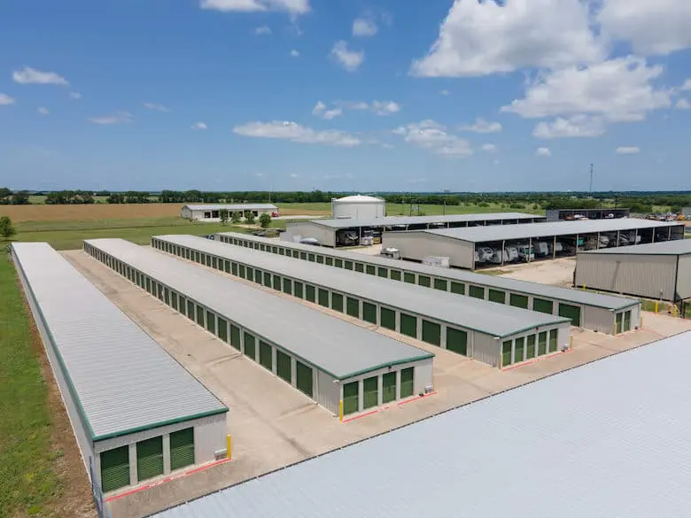 Aerial view of a large storage facility with multiple long, white buildings with green doors, surrounded by fields and under a blue sky with scattered clouds.